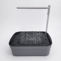 Grey bird bath with a dark slatted tray, water pouring from an overhead arm as a fine shower on a plain white background. 1