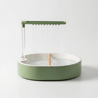 Green and white round bird bath on a plain background, water falling from a row of spouts onto a wooden perch. 1
