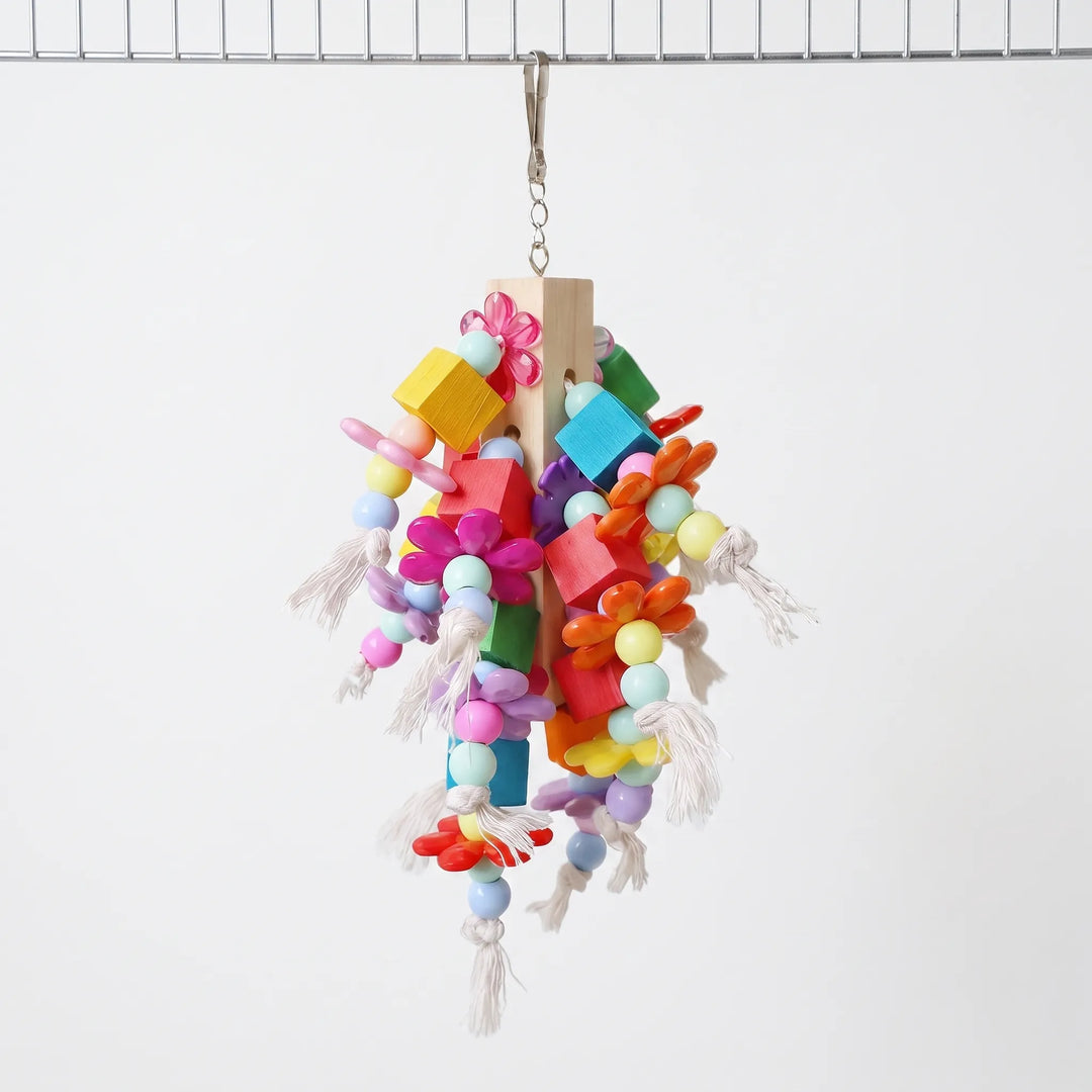 Bird toy hanging from a metal cage bar by a clip, with a wooden post, coloured blocks, flower beads and knotted cotton rope. 1