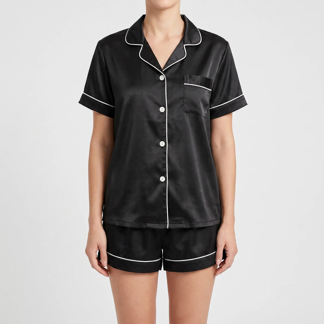 Front view of the black satin pyjama shirt and shorts with white piping, chest pocket and white buttons, worn by a model. 2