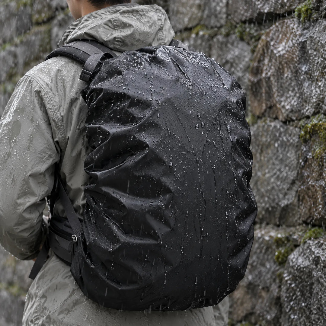 Hiker in a grey rain jacket seen from behind, wearing a black rucksack cover beaded with raindrops against a wet stone wall. 1