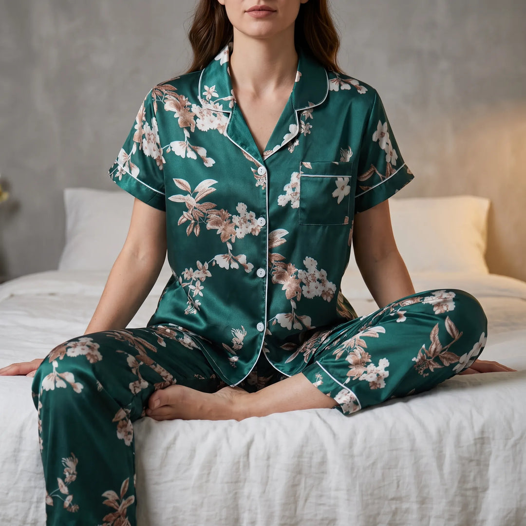 Model sitting cross-legged on a white-linen bed in the green satin-look short-sleeve pyjama set with floral print and white piping. 1
