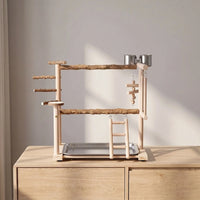 Wooden bird play stand on a pale wooden sideboard in soft daylight, seen from the front with perches, ladder and cups. 3