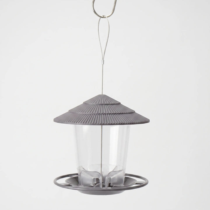 Grey feeder with ribbed tiered roof and clear seed tube, hanging from a steel wire loop and hook against a plain backdrop. 1