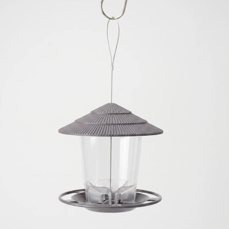 Grey feeder with ribbed tiered roof and clear seed tube, hanging from a steel wire loop and hook against a plain backdrop. 1