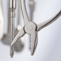 Close-up of the miniature silver pliers charm, showing the pivot and jaws, with other tool charms blurred behind. 3