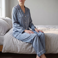 Woman sitting on the edge of a bed in blue satin pyjamas with a tonal heart pattern and white piping. 1