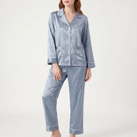 Model standing in blue satin pyjamas with heart jacquard pattern, front view showing buttons, two chest pockets and white piping. 2