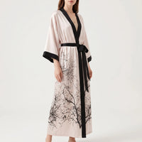 Three-quarter view of a model in the blush satin wrap robe, black trim and tied belt, branch print at the skirt. 2