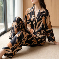 Model sitting on a beige carpet by a window in the black and tan abstract print satin pyjamas with orange piping. 1