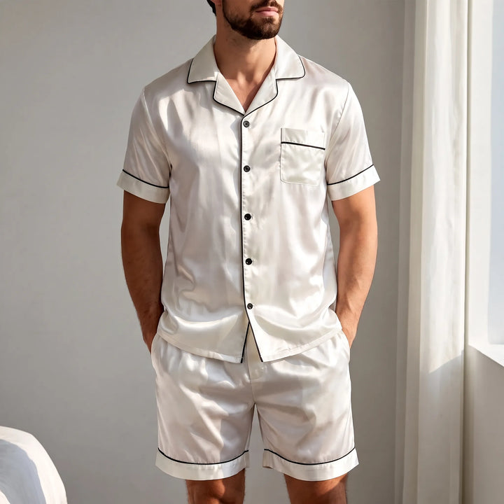 Model in white satin short-sleeve pyjama set with black piping stands relaxed by a bright window, hands in shorts pockets. 1