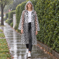 Model walking in the rain along a hedge-lined path, wearing the translucent hooded raincoat with a black leopard spot print. 1