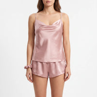 Front view of a model on a white background in the dusty pink satin cowl-neck cami and shorts, with a matching scrunchie on her wrist. 2