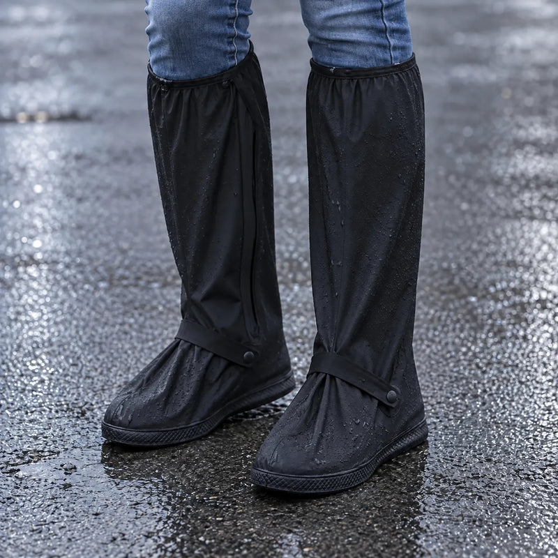 Black knee-high rain shoe covers with ankle straps worn over jeans on wet, rain-spotted asphalt, water beads on the fabric. 1