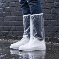 Clear knee-high overshoes with a white trim worn over white sneakers, standing in a rain puddle in front of a dark brick wall. 1