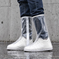 Side view of clear tall overshoes with a white strip along the leg, worn over white sneakers and dotted with raindrops against a concrete wall. 3