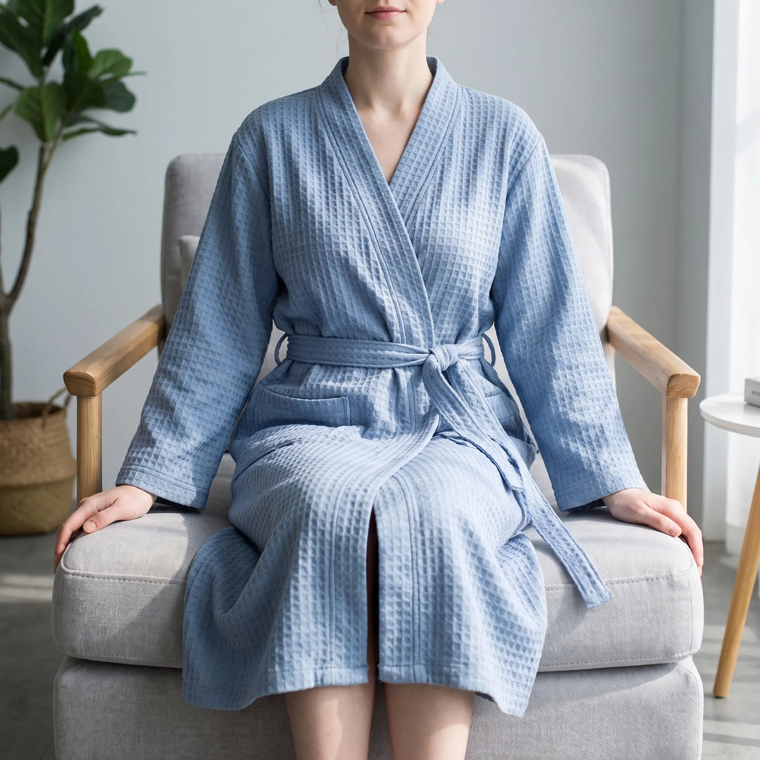 Woman sitting in a light armchair wearing the blue waffle-textured robe, belt tied at the waist, in a bright living room. 1