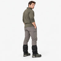 Man in a green long-sleeve top and grey trousers looks over his shoulder, wearing black gaiters over hiking boots. 4