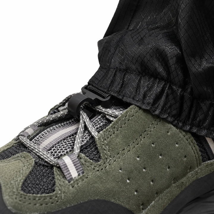 Close-up of the gaiter's lower hem with a plastic hook clipped to the laces of a green suede hiking shoe. 6