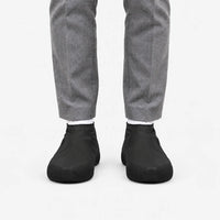 Front view of black rubber shoe covers worn with grey trousers and white socks, showing the textured grip at the toes. 4