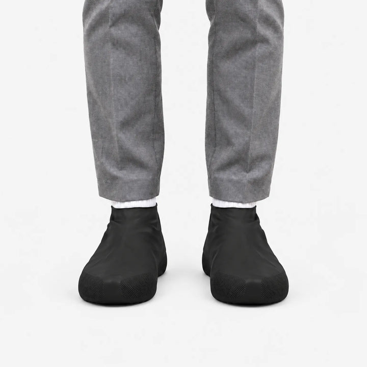 Front view of black rubber shoe covers worn with grey trousers and white socks, showing the textured grip at the toes. 4