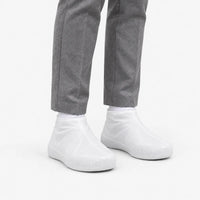 Angled view of translucent white rubber shoe covers worn with grey trousers, showing the textured toe area and snug ankle fit. 5