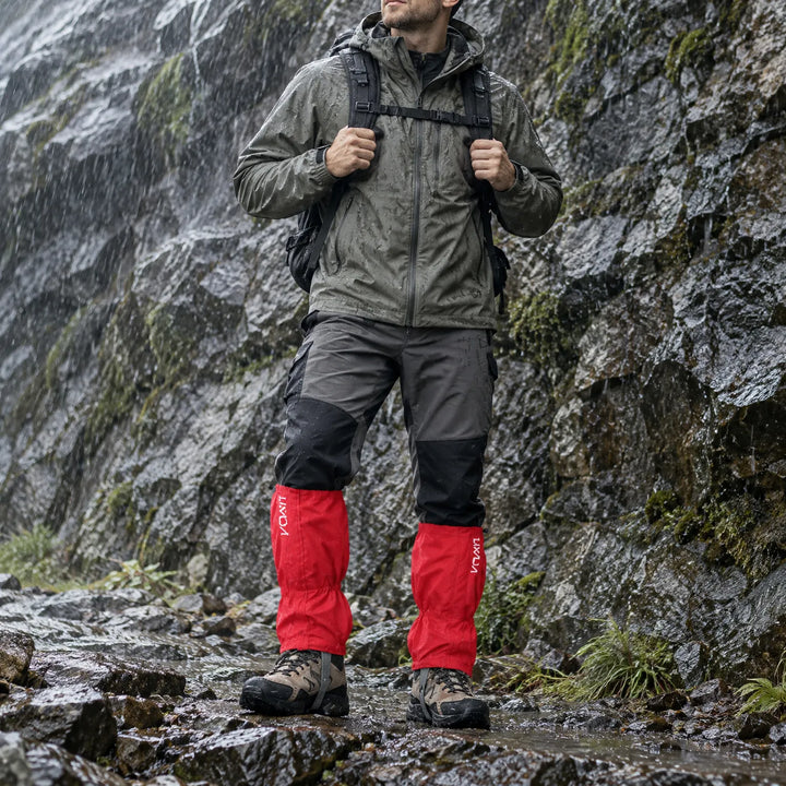Hiker in a rain jacket and backpack wears red gaiters over hiking boots while standing on a wet rocky trail beside a rock face. 1