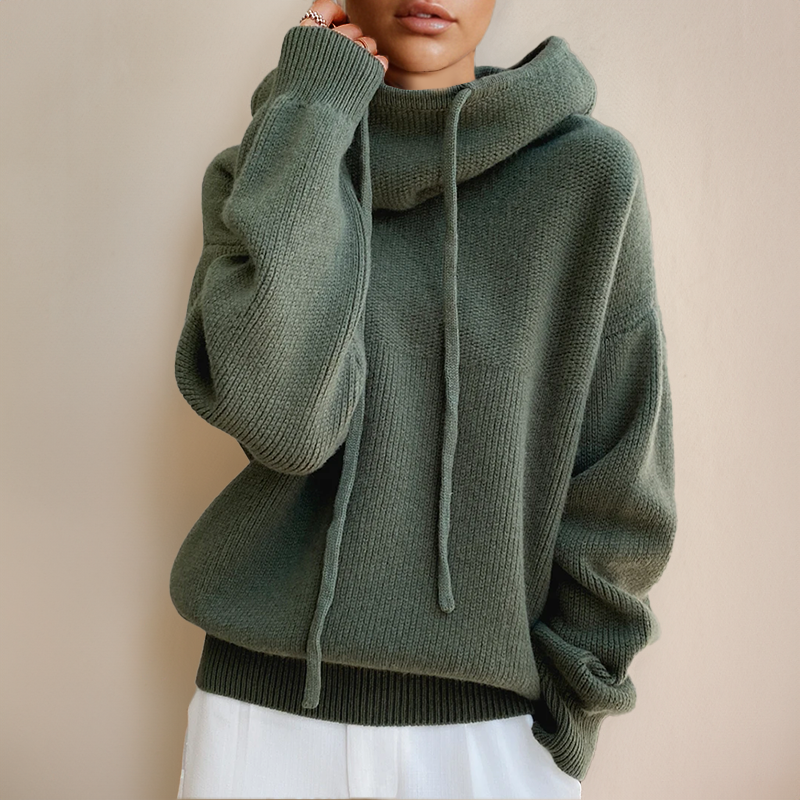 Close-up of a green chunky knit jumper with a slouchy cowl hood and long drawstrings, worn with white trousers 2